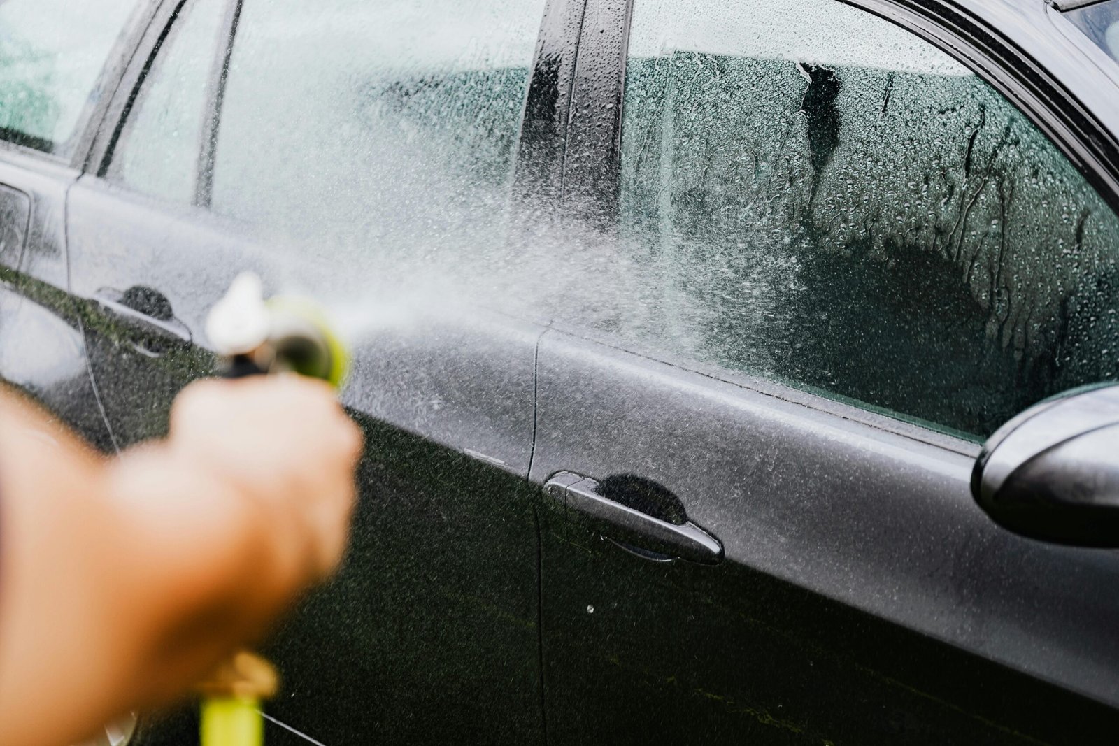 Услуги A car being washed with a hose outdoors. Water sprays create a cleaning effect on the vehicle.