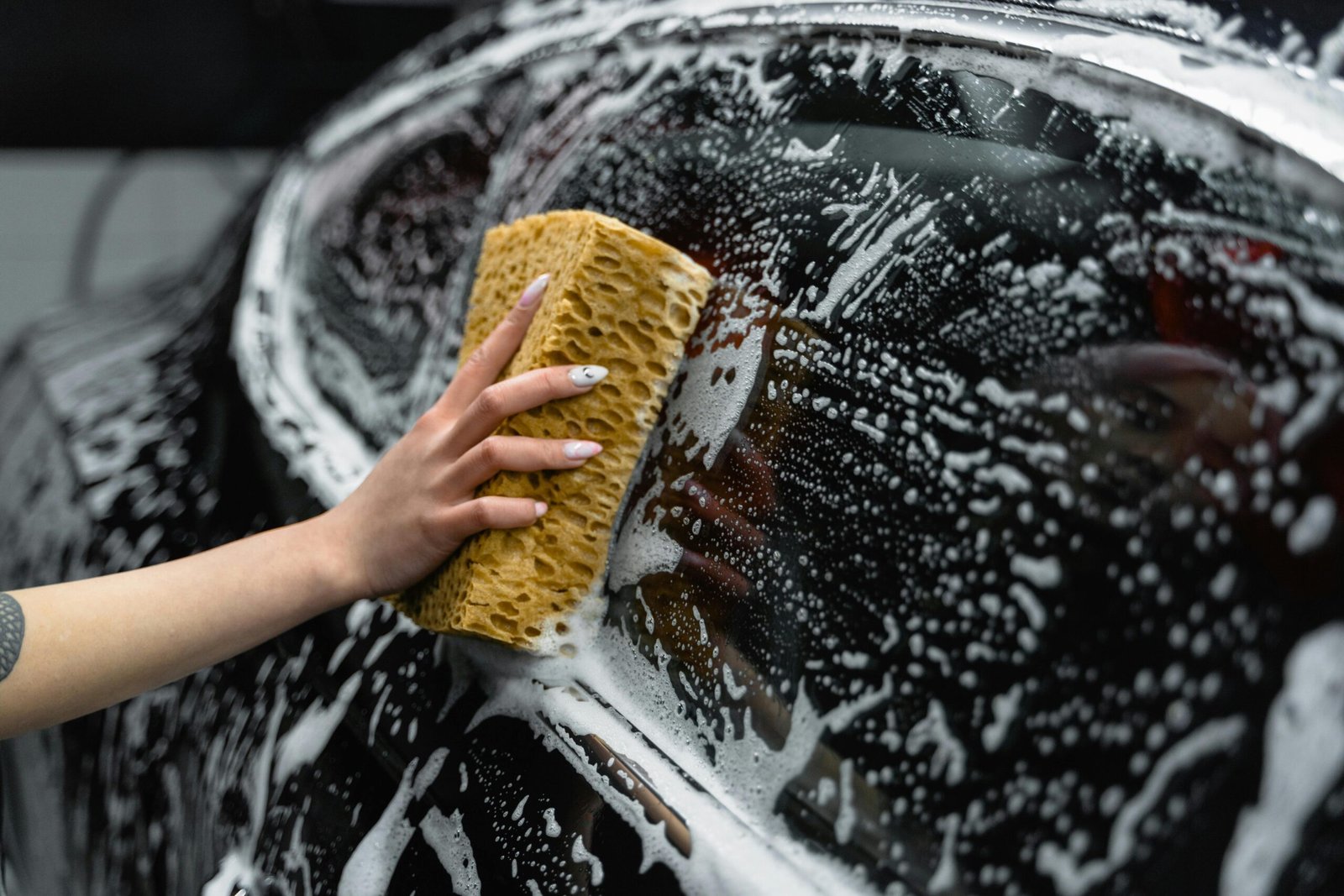 Услуги A hand scrubs a car window with a sponge, covered in soap and foam, at a car wash.
